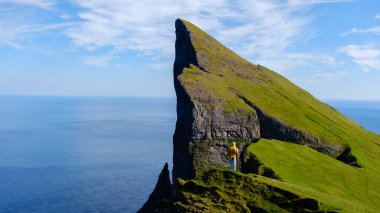 Yalnız bir gezgin, Faroe Adaları 'ndaki çarpıcı bir uçurumun tepesinde duruyor. Etrafı canlı yeşil çimenler ve sonsuz okyanus manzaralarıyla çevrili. Engebeli manzara dramatik kaya oluşumlarını ve sakin gökyüzünü sergiliyor..