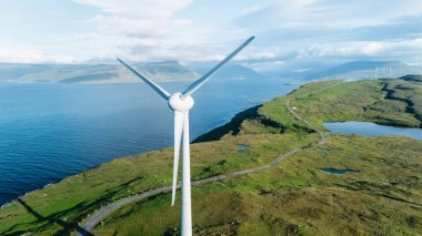 Bir rüzgar türbini, Faroe Adaları 'nın engebeli kıyı şeridi ve canlı yeşil tepelerinin üzerinde görkemli bir şekilde yükseliyor..