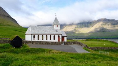 Yemyeşil tepelerin ortasında, dik çatılı, tuhaf beyaz bir kilise denizin kenarında gururla durur. Vidareidi Faroe Adaları 'nın arka planında kayan dağlar ve dramatik bulutlar
