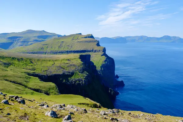 Görkemli uçurumlar, Faroe Adaları 'nın kıyı şeridi boyunca açık mavi bir gökyüzünün altında uzanır. Canlı yeşil tepeler ışıl ışıl Atlantik 'le buluşarak nefes kesici bir doğal manzara yaratıyor..