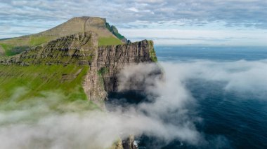 Faroe Adaları 'nın dramatik uçurumları, sislerin ve mavi suların zeminine karşı dimdik ayakta duruyor. Kaya yüzeyindeki yemyeşil şelaleler okyanus dalgalarına karşı çarpıcı bir kontrastlık yaratıyor..