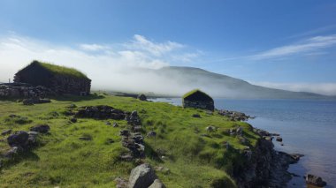 Faroe Adaları 'ndaki huzurlu kıyı manzarasında yemyeşil ve sisli tepelerle çevrili geleneksel çim çatılı kulübeler bulunur. Sakin sular manzaranın çarpıcı doğal güzelliğini yansıtır..