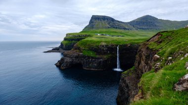 Nefes kesen bir şelale dalgalı uçurumlardan aşağı, Atlas Okyanusu 'nun dingin sularına akar, Faroe Adaları' ndaki canlı yeşil tepeler, Gasadalur köyü yakınlarındaki Mulafossur şelalesi