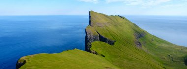 Bereketli yeşil kayalıklar, derin mavi okyanusun üzerinde Faroe Adaları 'nın büyüleyici doğal güzelliğini gözler önüne sererek dramatik bir şekilde yükselir. Güneş sıcak bir parıltı yayar, canlı manzarayı güçlendirir..