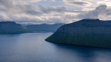 Faroe Adaları 'ndaki bir fiyordun sakin sularından nefes kesen uçurumlar dramatik bir şekilde yükselir. Yumuşak sabah ışığı manzarayı kaplar, yumuşak gölgeler bırakır ve engebeli adaları ortaya çıkarır..