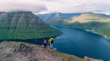 Bir çift, Faroe Adaları 'nın göz kamaştırıcı manzaralarını alan kayalık bir çıkıntıda el ele durur. Serin fiyortlar ve yemyeşil dağlar maceraları için mükemmel bir zemin oluşturur..