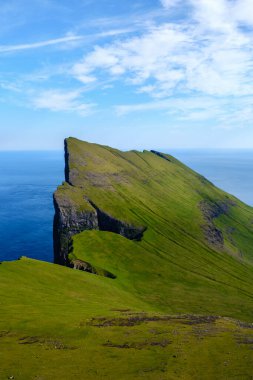 Mylingur uçurumu, parlak bir gökyüzünün altındaki engin mavi okyanusa bakan Faroe Adaları 'nın canlı yeşil manzarasından keskin bir şekilde yükselir. Doğanın güzelliği gözler önünde..
