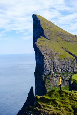Yalnız bir gezgin, Faroe Adaları 'ndaki Mylingur uçurumlarının kenarında, canlı yeşil manzaralar ve uçsuz bucaksız mavi bir okyanusla çevrili duruyor. Bu an, doğanın özünü yansıtıyor, majesteleri..