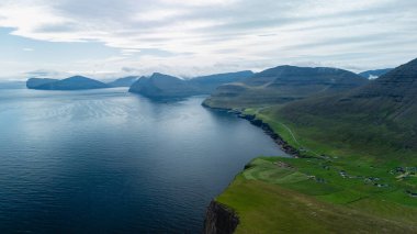 Faroe Adaları 'nın engebeli güzelliğini keşfedin. Bu çarpıcı hava manzarası dramatik uçurumlar, yemyeşil tepeler ve yumuşak mavi gökyüzünün altında sakin sular sergiliyor. Mükemmel bir yaz kaçamağı..