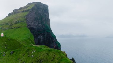 Yalnız bir yürüyüşçü, Faroe Adaları 'nın engebeli kıyı şeridine ve Kuzey Atlantik sularına bakan dramatik bir uçurum kenarında muzaffer bir şekilde duruyor. Kallur Deniz Feneri Kalsoy Adası