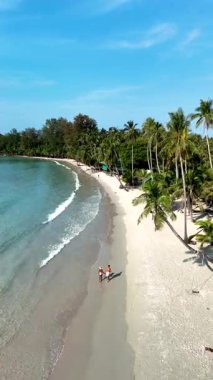 Tayland 'daki Koh Kood Adası' nın çarpıcı beyaz kumlu plajlarını keşfedin. İki insan kıyı boyunca yürür, bereketli palmiyeler ve berrak mavi sularla çevrili, canlı bir gökyüzünün altında..