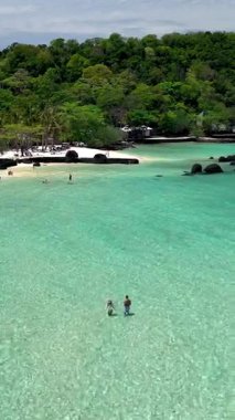 Koh Kham Adası nefes kesici turkuaz sulara ve rahatlama ve macera için ideal el değmemiş beyaz kumlu plajlara sahiptir. Birkaç beyaz erkek ve Asyalı kadın tatilde.