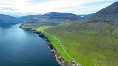 Açık mavi gökyüzünün altında Faroe Adaları 'nın çarpıcı sahil uçurumlarını ve yuvarlanan yeşil tepelerini keşfedin. Huzurlu sular, dingin bir atmosfer yaratarak, hareketli manzarayı yansıtıyor..