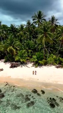 Discover the stunning shoreline of Koh Kood Island, Thailand, where two visitors stroll along the sandy beach surrounded by vibrant palm trees and clear turquoise waters under a dramatic sky.