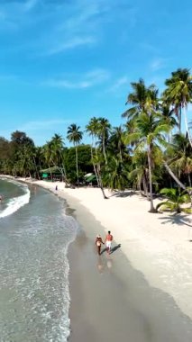 Relaxing on the pristine beaches of Koh Kood Island, Thailand, where soft sand meets crystal-clear water. A couple of sun seekers stroll along the shoreline, surrounded by lush palm trees