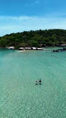 Crystal clear waters invite visitors to explore the tranquil shores of Koh Kham Island in Thailand. Lush greenery surrounds a pristine beach, a couple of caucasian men and Asian women on vacation