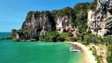Beautiful turquoise waters meet rugged cliffs in Krabi, Thailand. Longtail boats rest on the sandy shore, inviting visitors to explore this tropical paradise under the bright sun. Tonsai beach