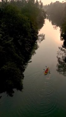 Paddling through the peaceful river of Chumphon, Thailand at dawn, this kayaker enjoys the natural beauty of lush greenery reflecting on calm waters. 