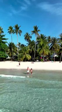A couple of men and women wade through the crystal-clear waters of Koh Kood Island, surrounded by tall palm trees and white sandy beaches. A perfect tropical getaway awaits on this beautiful island.
