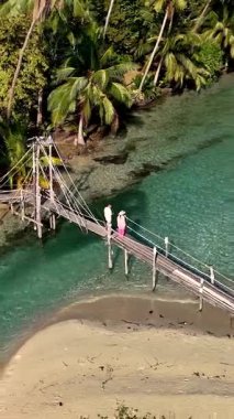 A couple stands on a wooden bridge over clear turquoise water in Koh Kood, Thailand. The picturesque landscape features vibrant palm trees and serene surroundings, perfect for a romantic getaway.