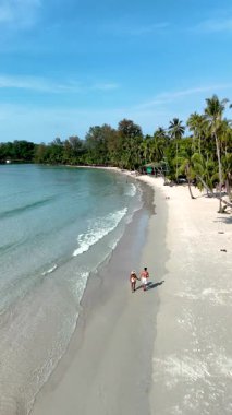 Experience the beauty of Koh Kood Island in Thailand, where soft sandy beaches meet tranquil waters. Two people stroll along the shore, surrounded by lush greenery and clear blue skies.