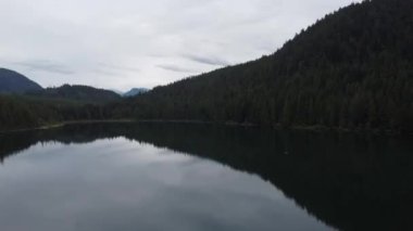 Gölün üzerinden uç bulutlu gökyüzünü ve etrafındaki yeşil ladin ormanını yansıt