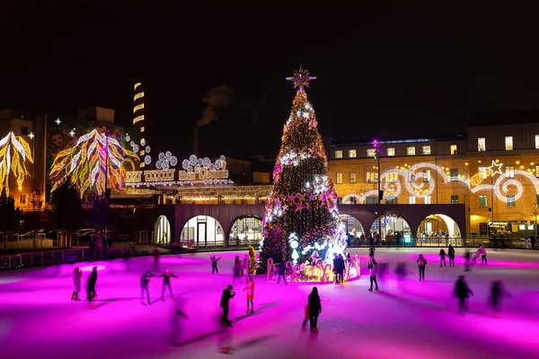 A Christmas tree in the middle of the ice rink. People skate around the rink. 