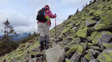 Bir kadın yürüyüş yaparken kayaların üzerinden dağa tırmanır. Yürüyüş sopaları kullanıyor.