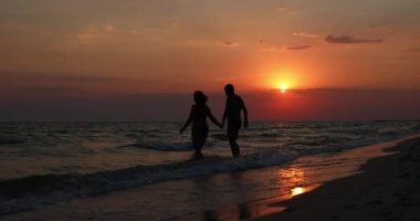 Bir adam ve bir kadın, güzel bir günbatımının arka planında el ele tutuşarak denize doğru yürüyorlar..