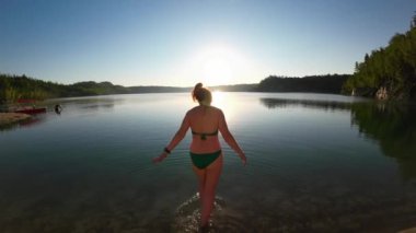 Güneşli bir yaz sabahının erken saatlerinde bir kadın gölün berrak sularında yürür. 