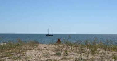 Güneşli bir öğleden sonra, bir kadın deniz manzaralı bir tepeye doğru yürür ve uzaktaki bir yatla..
