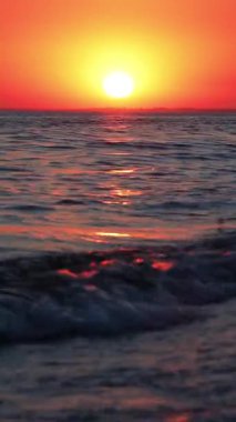 Denizin dalgaları kıyıda kırılır. Güneş ufuktan batıyor, gökyüzünü turuncuya çeviriyor. Bir martı manzaranın arka planında uçar..