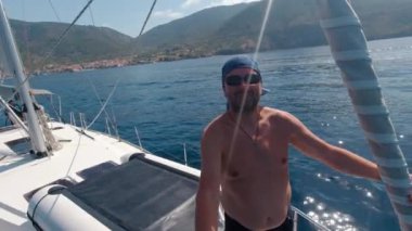 Hareket halindeki bir yatın pruvasında bir adam kameraya çevresini gösteriyor..