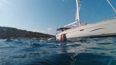 A woman enjoys swimming in the sea with a yacht in the background on a sunny summer day. Over and under.