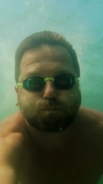 Gözlüklü bir adam suyun altında dalıp yüzüyor. Dikey Video