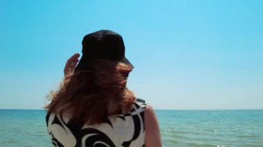 The woman in a bucket hat turns toward the camera in slow motion, set against the backdrop of a bright summer day by the sea, highlighting her sunny beach look