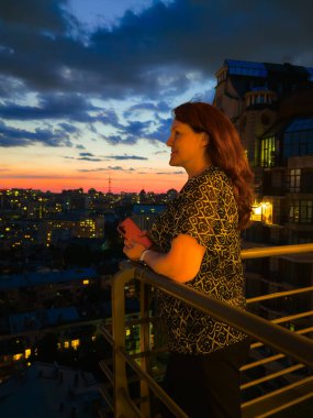 Bir kadın balkonda duruyor, akşam şehrini gün batımının arka planına bakıyor.