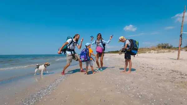 Güneşli bir plajda, bir aile sırt çantaları ve yanlarında köpekleri ile kıyı yürüyüşüne çıkmaya hazırlanıyor, önlerindeki macerayı sabırsızlıkla bekliyorlar.