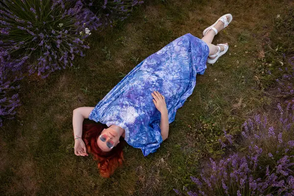 Mavi elbiseli ve pembe güneş gözlüklü bir kadın lavanta çalılarının arasında yerde yatıyor.