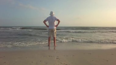 Kova şapkalı adam kıyı şeridinde rahat duruyor, dalgalar nazikçe ayaklarına vuruyor, yaz akşamının sıcağının tadını çıkarıyor.