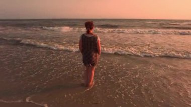 Sıcak bir yaz akşamında, kadın sörfte duruyor, günbatımının dingin güzelliğinin ve ayaklarına vuran nazik dalgaların tadını çıkarıyor.