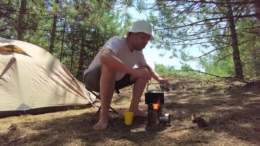 Adam çaydanlıktan bir bardağa sıcak çay döker, onu ateşten yeni çıkarmış, güneşli çam ormanında çadırının yanında otururken.