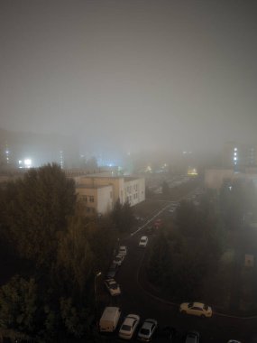 Büyüleyici bir şehir manzarası gece sisi ile dönüşür, pencerelerden ve sokak lambalarından gelen ışıklar sislerin arasından sıcak bir parıltı yayarak, sıcak bir sonbahar atmosferi yaratır.
