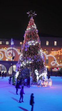 Renkli ışıkları olan neşeli bir Noel ağacı, akşamın tadını çıkaran patencilerle çevrili, parlayan bir buz pateni pistinde dimdik ayakta duruyor. Yakınlarda, tatil süslemeleriyle parıldayan bir bina.. 