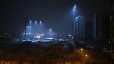 Endüstriyel inşaat alanı ruhani mavi spot ışıkları ile yıkandı. Gece sisini delip geçti. Sıcak sokak lambaları, yüksek vinçlerin altındaki zemin seviyesini aydınlatırken çarpıcı ışık huzmeleri oluşturdu.