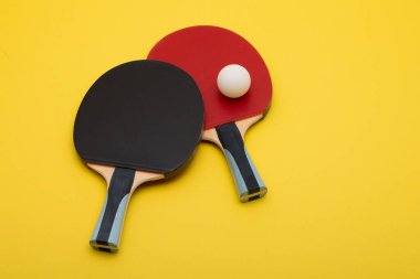 Kırmızı ve siyah kauçuk yüzeyli iki profesyonel pinpon raketinin yer aldığı şık kompozisyon, canlı sarı arka planda düzenlenmiş beyaz rekabet topu eşliğinde.