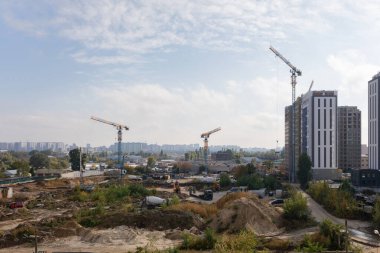 Üç vinç ve çeşitli inşaat araçlarının çalıştığı bir inşaat alanı. Ön planda, bitmemiş bir binanın yanında uzanan kum yığını devam eden ilerlemeyi vurguluyor.