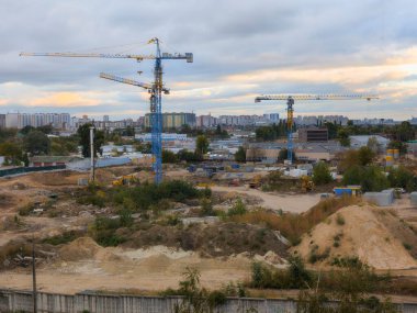 Hareketli bir inşaat sahasında, yeni istiflenmiş toprak ve ufuktaki yerleşim alanlarının ufkuyla çevrili çeşitli vinçler ve ağır makineler sergileniyor.