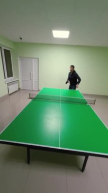 Yeşil masa tenisi ekipmanlarıyla kapalı masa tenisi tesisinde heyecan verici bir maça hazır bir oyuncu var. Açık yeşil duvarları olan temiz, iyi ışıklandırılmış bir oda eğlence sporları ve fitness aktiviteleri için mükemmel. Dikey Video