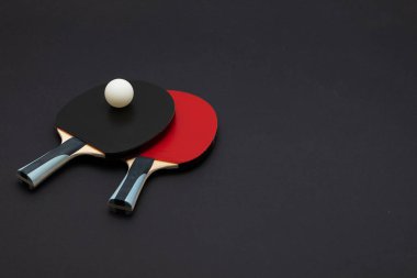 Karşıt siyah ve kırmızı kauçuk yüzeyler, tahta saplar ve koyu renk arka plana karşı rekabet topunun yer aldığı iki profesyonel masa tenisi raketinin dramatik çekimleri.